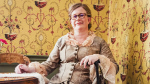 Piia Vähäsalo on panostanut kauppahuoneen sisustukseen. Piia Vähäsalo istuu 1800-luvun mekkoon pukeutuneena, kävelykeppiin nojatessa, kauppahuoneen kahvilan puolella.