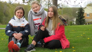 Två av de här tjejerna funderar på att läsa medicin och en är intresserad av biologi och modersmål. Men första etappen efter avslutad grundskola är Ekenäs gymnasium. Från vänster Wilma Lindholm, Milla Hyvönen och Emilia Kytöpuro. Tre tonåringar (flickor) sitter på en grön gräsmatta med några gula maskrosor. Ett träd och ett hus långt i bakgrunden.