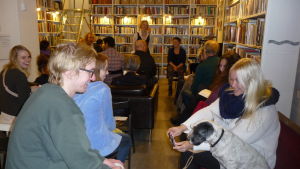 I väntan på att kvällens litterära afton ska börja. Publik i en bokhandel. De sitter och väntar på att en föreställning ska börja. En hund väcker publikens förtjusning.