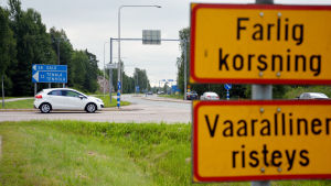 En vit bil står i en korsning. I förgrunden syns ett trafikmärke där det står farlig korsning.