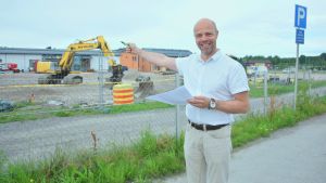 Markus Moisio vid skolbygget i fjol. Man i vit kragskjorta framför byggarbetsplats på stor grusplan med stängsel omkring. Mannen pekar mot arbetsplatsen.