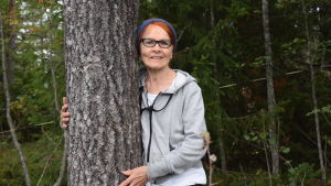 Marjatta Lyytikäinen är glad över att få bo mitt i skogen. Hennes tomt gränsar till en planerad odlingslott, gemensam för bostadsområdets invånare. Just nu är hon också ensam herre på (trädgårds)täppan. En kvinna med rött hår, glasögön och grå munkjacka kramar om en tall.