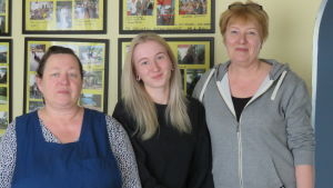 Vänstugans mathjälpsteam just nu: Annika Söderlund lagar mat, Janina Gabrielsson packar ihop mat och Vänstugans verksamhetsledare Ann-Maj Gabrielsson håller kontakt med hjälpbehövande och stamgäster samtidigt som hon deltar i matutdelningen. Tre kvinnor står framför en gul vägg med många fotografier.