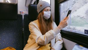 Kvinna i grå mössa och beige jacka och munskydd åker tåg, tittar på sin mobiltelefon.