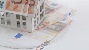 Hangö stads bolag får mera tid på sig att betala tillbaka lånen. En temabild av ett miniatyrhus vars tak är gjort av femtioeuros sedlar. Över huset ligger en glaskupa.