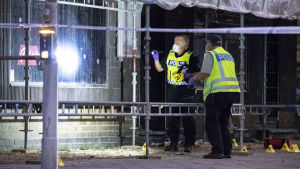 Två poliser undersöker skotthål i ett fönster. Framför fönstret står också byggställningar.