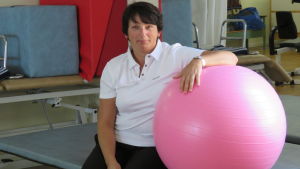 En kvinna med mörkt kort hår och vit skjorta sitter och håller om en stor ljusröd jumppaboll. I bakgrunden stor spegel och stora röda och blå plastmadrasser som används i olika terapier.