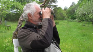 En stol och en kikare är allt du behöver för att komma närmare naturen i din omgivning. Dick Forsman sitter i sin trädgård och spanar med kikare.