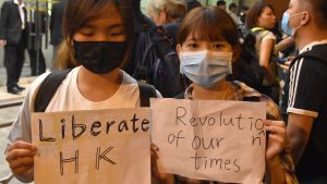 De här unga kvinnorna skulle begå ett allvarligt brott i dagens Hongkong. Deras slogan ”Befria Hongkong, vår tids revolution” har förklarats bryta mot den nya säkerhetslagen. Två kvinnor i munskydd håller upp skyltar där det på engelska står "berfria Hongkong" och "vår tids revolution"