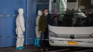 WHO-delegationen anlände till en avspärrad del av Wuhans flygplats på torsdagen. De sattes på en buss och fördes till ett hotell där de ska vara två veckor i karantän. Personer med munskydd omgivna av vakter i vita heltäckande skyddsdräkter stiger på en vit buss.