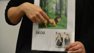 Sandra håller i ett ark med ett lejon på och där det står skog. Sandra visar tecknet för skog.