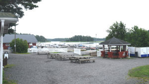 Sommartid ligger många båtar vid Sommaröstrand. En hamn, i förgrunden syns bord och bänkar och i bakgrunden småbåtar som ligger vid bryggor nära stranden. I bakgrunden öar och vatten.