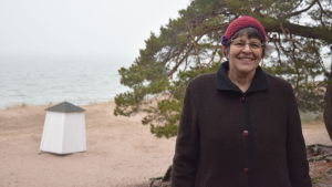 Astrid Lindström, ordförande för Hangö miljöförening. En kvinna iklädd vinterkläder poserar framför en strand med tallar och sanddyner.