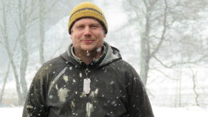 Vinterns enda rejäla snöfall i Ingå kom i början av april när de flesta ville fortsätta njuta av den tidiga våren. Gustav Hildén är på gott humör trots snön. Han har fått många sommarjobbsansökningar av traktens ungdomar. En man i gul mössa och grå jacka står på en åker i ymnigt snöfall.