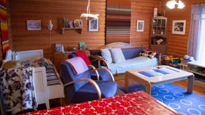 Det lönar sig att plocka bort värdeföremål ur sommarstugan innan någon annan gör det. Vardagsrummet i en sommarstuga med stockväggar, utdragbara bäddsoffor, en blå fåtölj och ett matbord med en blommig bordduk.