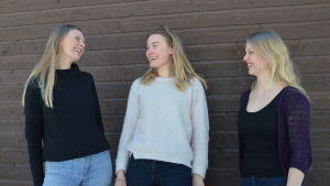Tre kvinnor står framför en trävägg. De tittar på varandra och skrattar. De är utomhus. 