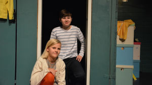 Alma Nordqvist och Sebastian Rönnholm. Två barn sitter på huk på en teaterscen. I bakgrunden färgglada lådor.