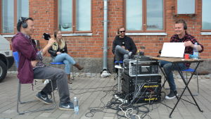 Kulturjournalisten Tomas Jansson, Viper Arms basist Soffe, Sivujuttus sångare Martin Laine och redaktören Joakim Rundt i direktsändning om rockmusiken i Åbo. Kulturjournalisten Tomas Jansson, Viper Arms basist Soffe, Sivujuttus sångare Martin Laine och redaktören Joakim Rundt sitter på stolar framför en tegelvägg i en direktsändning om rockmusiken i Åbo.