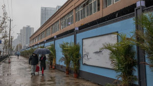 Den nedre våningen i Wuhanmarknaden där det handlades med färskvaror och levande djur är stängd sedan ett år. En lång tvåvåningsbyggnad med ljusblå skivor som avspärrning. Några människor promenerar förbi i regnet.
