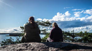 Ersätt skrollandet med att till exempel träffa en vän ute i naturen. Kaksi naista istuu korkealla kalliolla ja katselee kohti merta maisemaa.