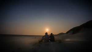 kaksi hahmoa istuvat huput päässä hiekalla auringon laskiessa