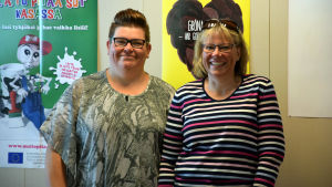 Linda Hellman och Veronica Forsstedt hjälper till med serveringen i skolmatsalen. Två kvinnor står vid en vägg och skrattar mot kameran.