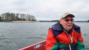 Paavo Paju är inte fiskare men kommer från ett familj av flera generationer fiskare. Nu jobbar han för yrkesfiskarna genom att bland annat vara ordförande för Västra Nylands fiskeområde. En bild på en man i röd och blå flytvästjacka och med solbrillor och keps. I bakgrunden syns hav och öar. På en av öarna bor en skarvkoloni och därför är många träd döda.
