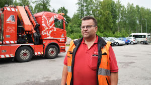 Ulf Österman driver Östermans alltrans i Karis och har ungefär tio anställda. En man står i förgrunden iklädd röd t-skjorta och reflexväst. Han har glasögon. I bakgrunden syns en lastbil som kan bärga bilar och bakom den skog.
