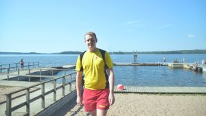 Kasper Rehn har kunnat leverera goda nyheter om algläget till badgästerna. Badvakt i gul t-skjorta och röda shorts vid sandstrand. I bakgrunden en brygga.
