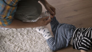 I dagens läge betalas hemvårdsstöd ut för barn under tre år som inte har en kommunal dagvårdsplats. En stor majoritet av dem som lyfter hemvårdsstöd är kvinnor. Vanhempi laittaa sukkaa lapsen jalkaan.