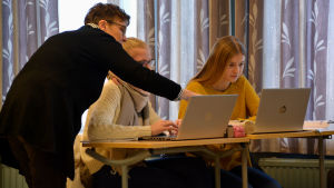 Gymnasieläraren Camilla Niiranen hjälper. Läraren hjälper två flickor, sträcker fram sin hand och pekar på dataskärmen.
