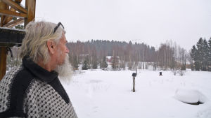 Bakom träden kan man skymta grannen Seppo Vuoksenturjas hus och ladugården där skyddshemmets djur också kan vara. En äldre man ser ut över ett snötäckt landskap.