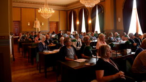 Öppningsseminariet hölls i fullmäktigesalen i stadshuset. En fullmäktigesal är full med människor som sitter och lyssnar på ett seminarium.