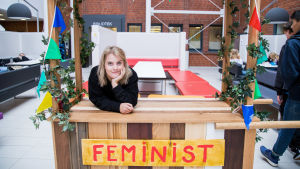 Ulrikke Falch nojaa pienen puisen kioskin tiskiin. Kioskin edessä on kyltti, jossa lukee feminist.
