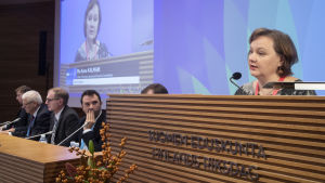 Anne Kalmari (c) på europeiskt möte för jordbrukspolitiker. Anne Kalmari i talarstol och på storskärm i riksdagens mötessal