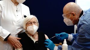 Edith Kwoizolla vaccinerades i Tyskland i går. Äldre kvinna med vitt hår, munskydd och glasögon får vaccin av man i blå skyddsdräkt.
