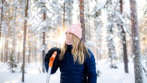 Nainen seisoo lumisessa metsässä nojaten lumilapioon.