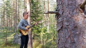 Bakom huset börjar skogen. I förgrunden en granstamm, i bakgrunden kvinna med gitarr.