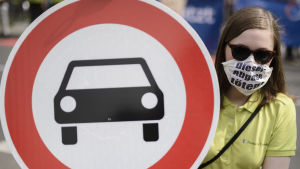Protest mot dieselavgaser. En kvinna med en ansiktsmask där det står "diesel-avgas dödar" håller upp ett trafikmärke med en bil på.