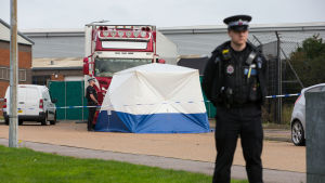 Polisavspärrningar runt långtradaren där de döda migranterna upptäcktes i Essex i oktober 2019. Till höger en polis. I bakgrunden ett blåvitt tält och bakom det en röd lastbil.