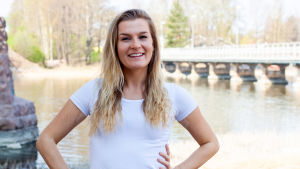 Som livsstilsrådgivare hjälper Corinne Grönholm sina kunder att genomföra förändringar i livet. En kvinna med långt blont hår och vit t-shirt står och ler. I bakgrunden syns en sjö och en bro.