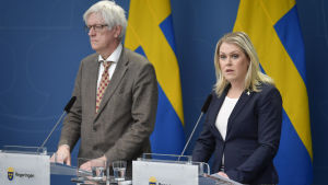 Folkhälsomyndighetens Johan Carlson och socialminister Lena Hallengren försäkrar att Sverige får vaccindoserna lika snabbt som andra EU-länder. En man i grått hår och grå kostym med kravatt med röda inslag står bakom ett podium som det står regeringen på och där det är emblemet tre kronor på. Bredvid honom en kvinna med axellångt ljust hår och blå kavaj o randig kjol, bakom likadant podium.