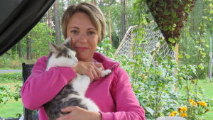 Jenny Grabber med familjens minsting, katten Zelda. En kvinna sitter ute, under ett partytält/paviljong och håller en katt i famnen. Sommar, mycket blommor och grönt.