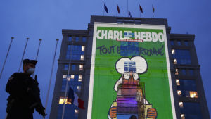 En byggnad där ett omslag på satirtidningen Charlie Hebdo projicerades på en vägg till minne av Paty. Bild på polis med munskydd utanför byggnad där en stor bild av satirtidningen Charlie Hebdos uppslag projiceras på fasaden till minne av Samuel Paty.