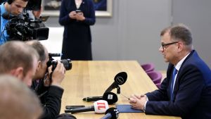 Juha Sipilä meddelar muntert att regeringskrisen är avblåst. Beskedet kommer under en snabbt sammankallad presskonferens på Åbo flygfält. Juha Sipilä sitter vid ett bord. Fotografer på andra sidan bordet.