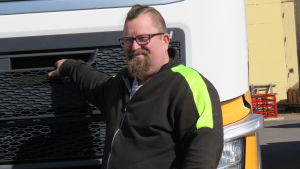 I vanliga fall brukar lastbils- och långtradarchaufförena hjälpa till inne i hallen med att lasta bilen. Nu får Magnus Forsman hållas inne i sin lastbilshytt förutom när han är utomhus, på behörigt avstånd från alla andra. Lunch och toalettbesök måste han fixa någon annanstans än på arbetsplatsen i Hangö. En man med kort och delvis snaggat hår, skägg och glasögon står framför en lastbilshytt. Han är klädd i arbetskläder i svart och neongult.
