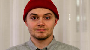 Christoffer Nyström arbetar på en guldsmedsaffär i Helsingfors. Porträtt på en ung man med skägg, en grå kragskjorta och en röd mössa på huvudet.