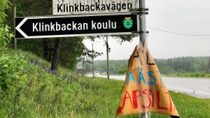 "Älä säästä lapsilta", på svenska "Spara inte på barnen", står det på ett regnpiskat plakat som dykt upp i närheten av Klinkbackan koulu. En vägskylt med finsk och svensk text. Nedanför vägnamnen står det på en svartvit skylt Klinkbackan koulu. Till höger löper en asfaltväg. Regnig väder. Grön försommarmiljö.