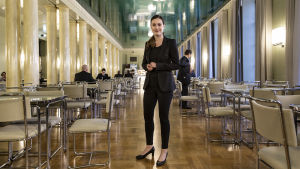 Sanna Marin poserar i riksdagens café. Sanna Marin, en ung kvinna i svart kavaj, svarta byxor och svarta högklackade skor står i en stor sal med många bord och stolar. Salen är kantad av pelare på ena sidan.