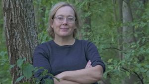 Valokuvaaja Maija Blåfield nojaa hyväntuulisena puunrunkoon kesäisessä metsikössä. 